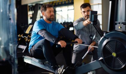 Mature athlete man using rowing machine during exercise class with personal trainer in a gym.