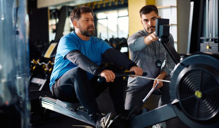 Mature athlete man using rowing machine during exercise class with personal trainer in a gym.